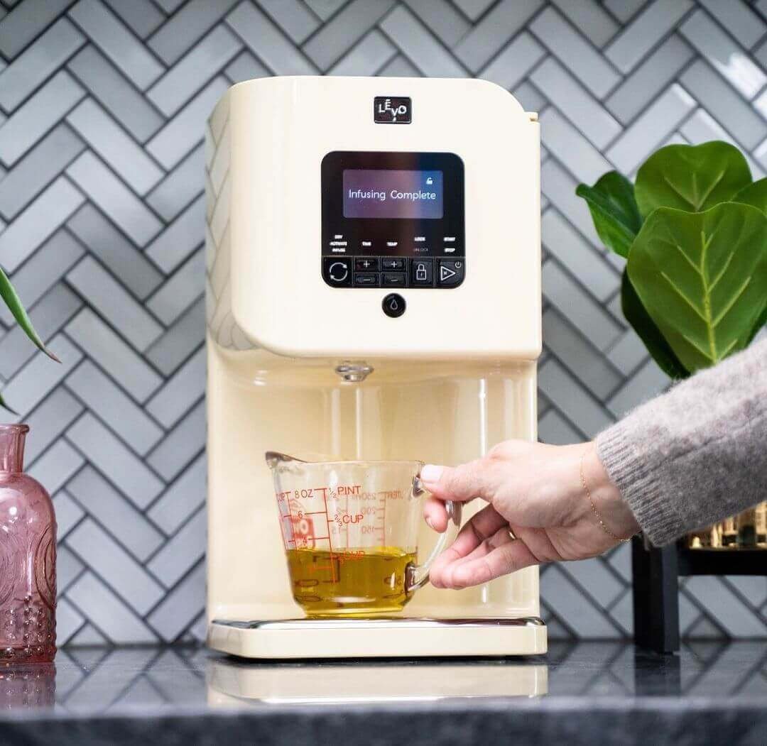Hand pouring golden cannabis-infused oil from a Levo brand countertop infusion machine into a measuring cup with red graduations