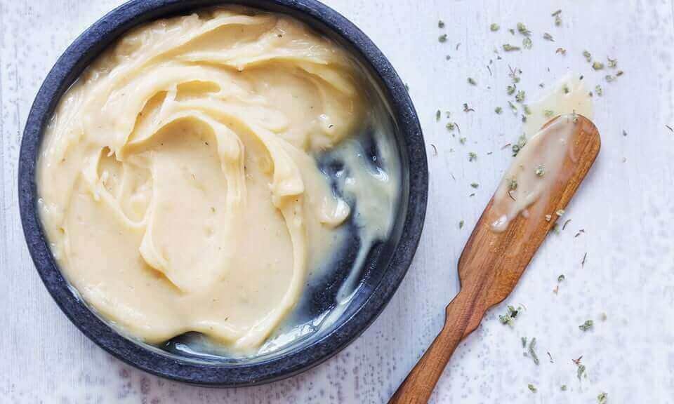 Smooth golden-yellow cannabutter in a dark ceramic bowl with a wooden spreader on a white wood surface, scattered with herb flecks