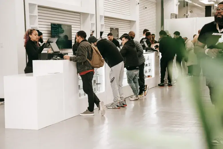 Busy urban dispensary with line of customers leaning over white display counters checking products