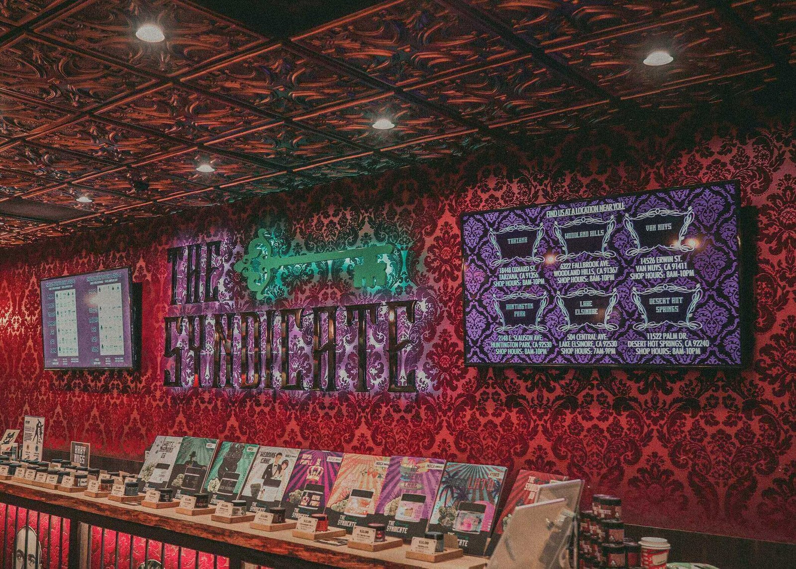 Dimly lit dispensary interior with red damask wallpaper and The Syndicate sign behind display cases