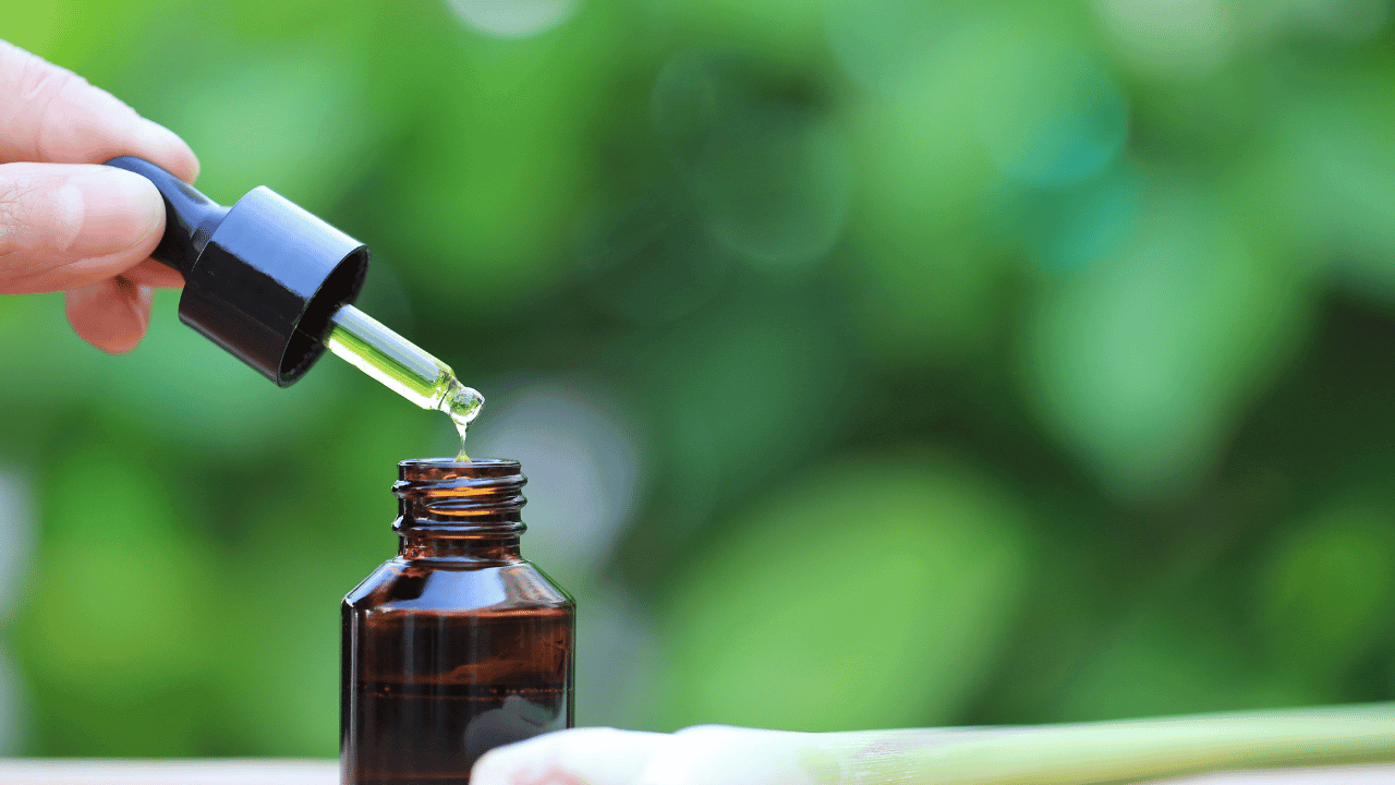 Hand dripping yellow tincture oil from black dropper into amber glass bottle outdoors with green foliage