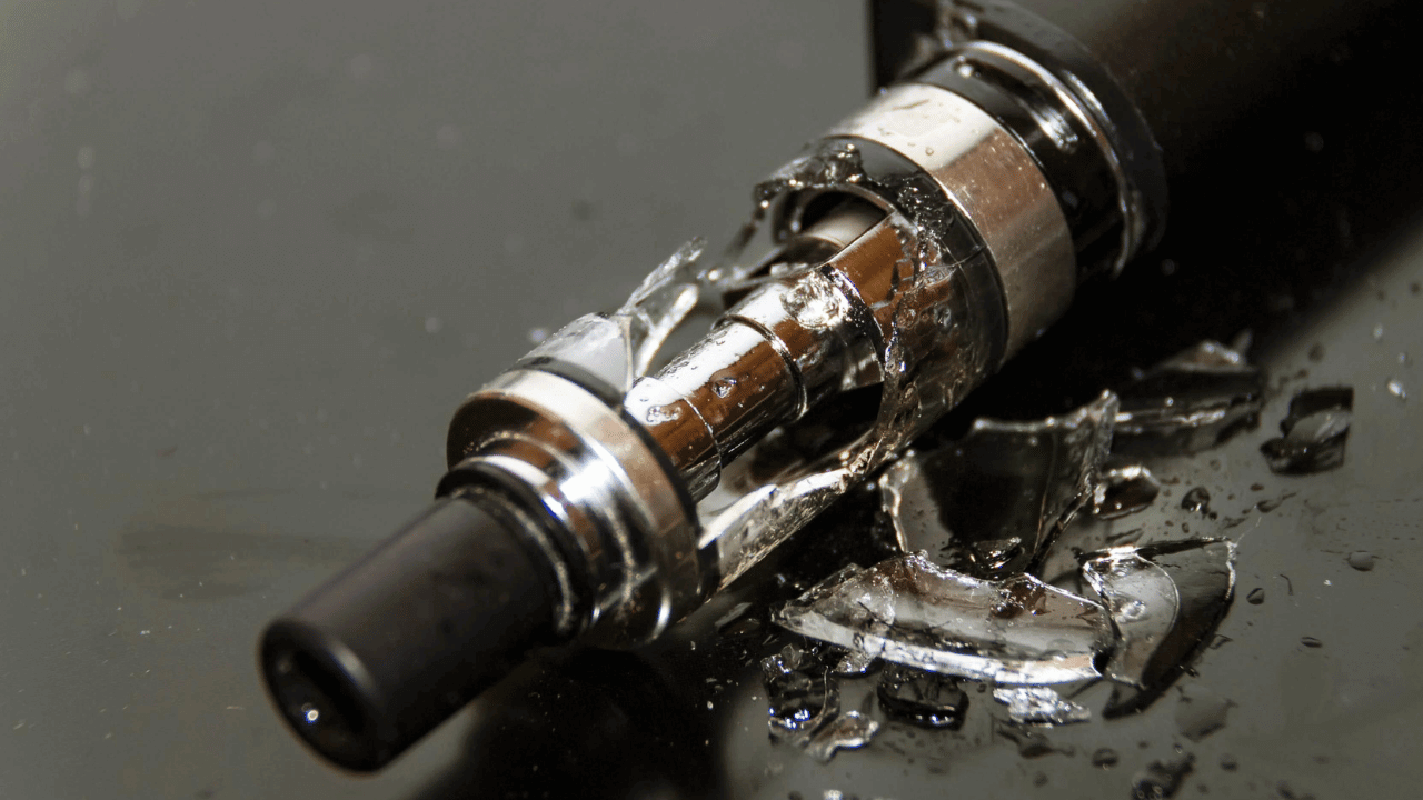 Broken vape pen cartridge with shattered glass scattered on dark reflective surface