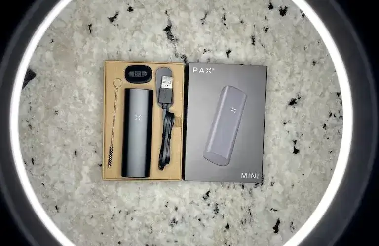Black Pax Mini vaporizer in retail packaging beside USB charging cable on speckled stone surface