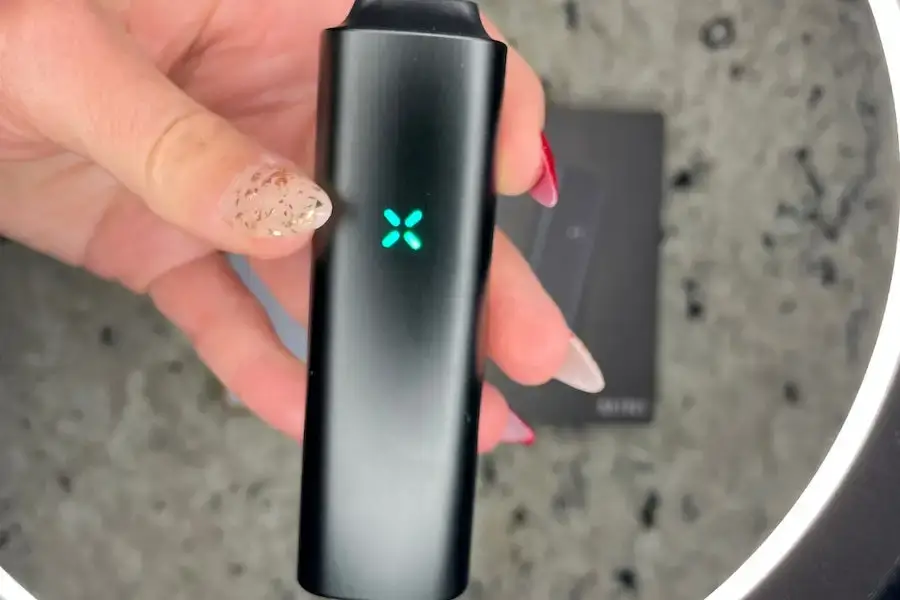 Hand holding a black Pax vaporizer device with glowing teal X-shaped LED indicator