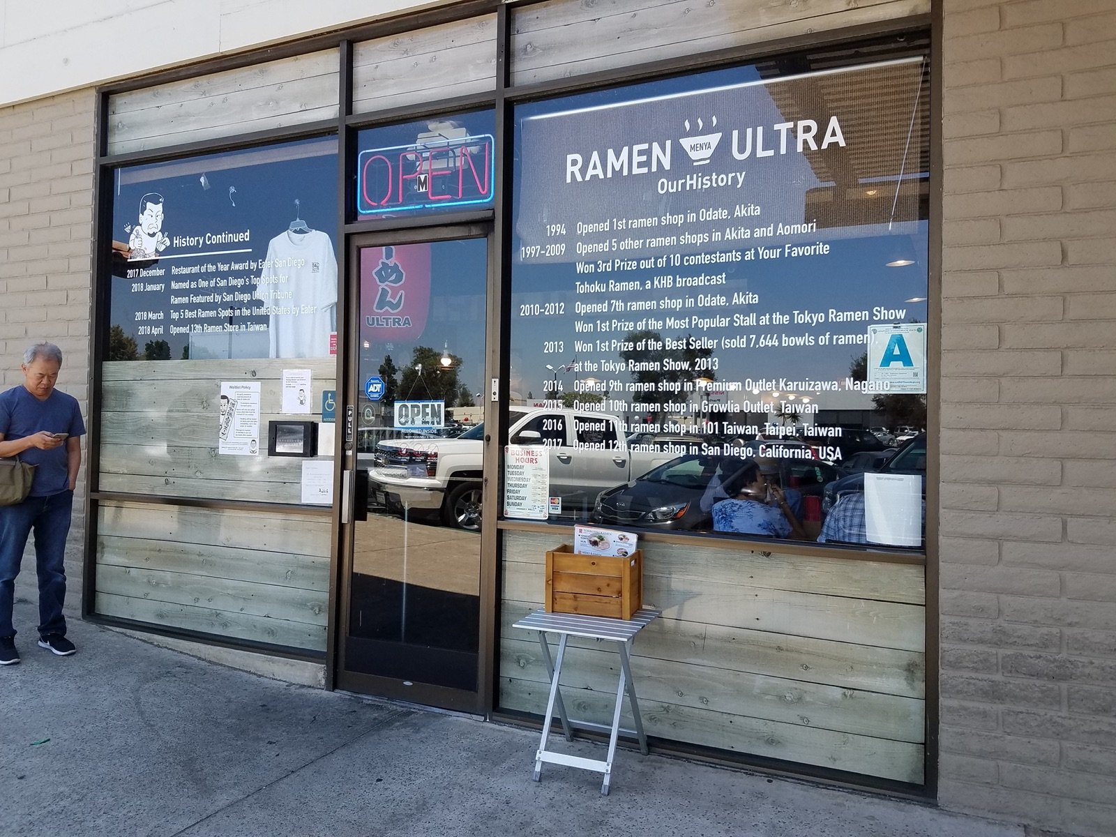 Exterior of Menya Ultra Ramen in Kearny Mesa San Diego with a history wall of awards and an OPEN sign visible through the storefront