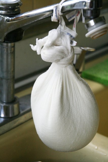 Cheesecloth bag tied to a kitchen faucet hanging over a sink, draining slowly by gravity without being squeezed