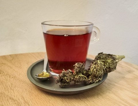 Cannabis-infused beverage in a clear glass beside dried cannabis flower on a wood table, representative of the cannabis beverage category Ayrloom produces