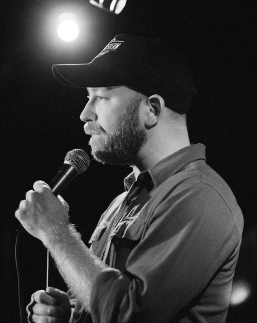 Stand-up comedian Kyle Kinane mid-set with the microphone, the bearded working-class storyteller of the American strip-mall smoke sesh.