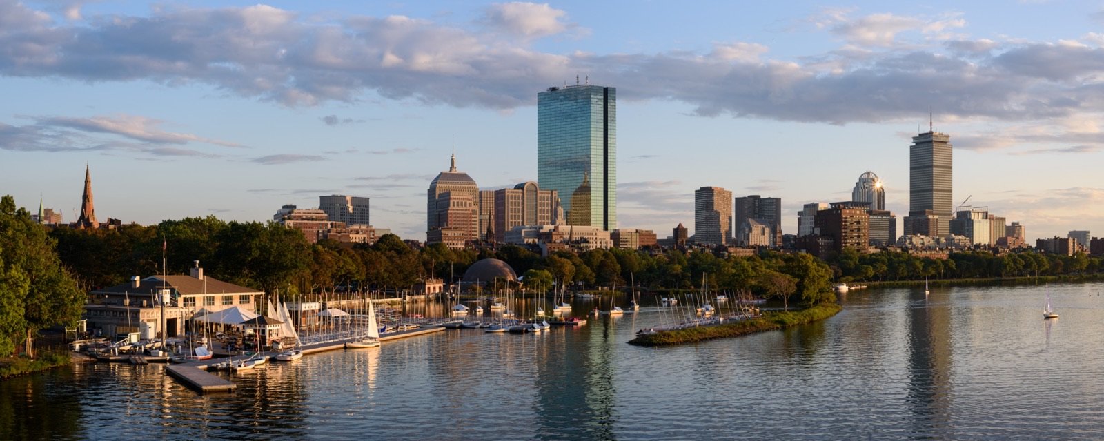 Boston skyline reflected over the Charles River from the Longfellow Bridge with the John Hancock Tower and Prudential Building anchoring the Back Bay, representing the Massachusetts adult-use cannabis market that launched on November 20, 2018