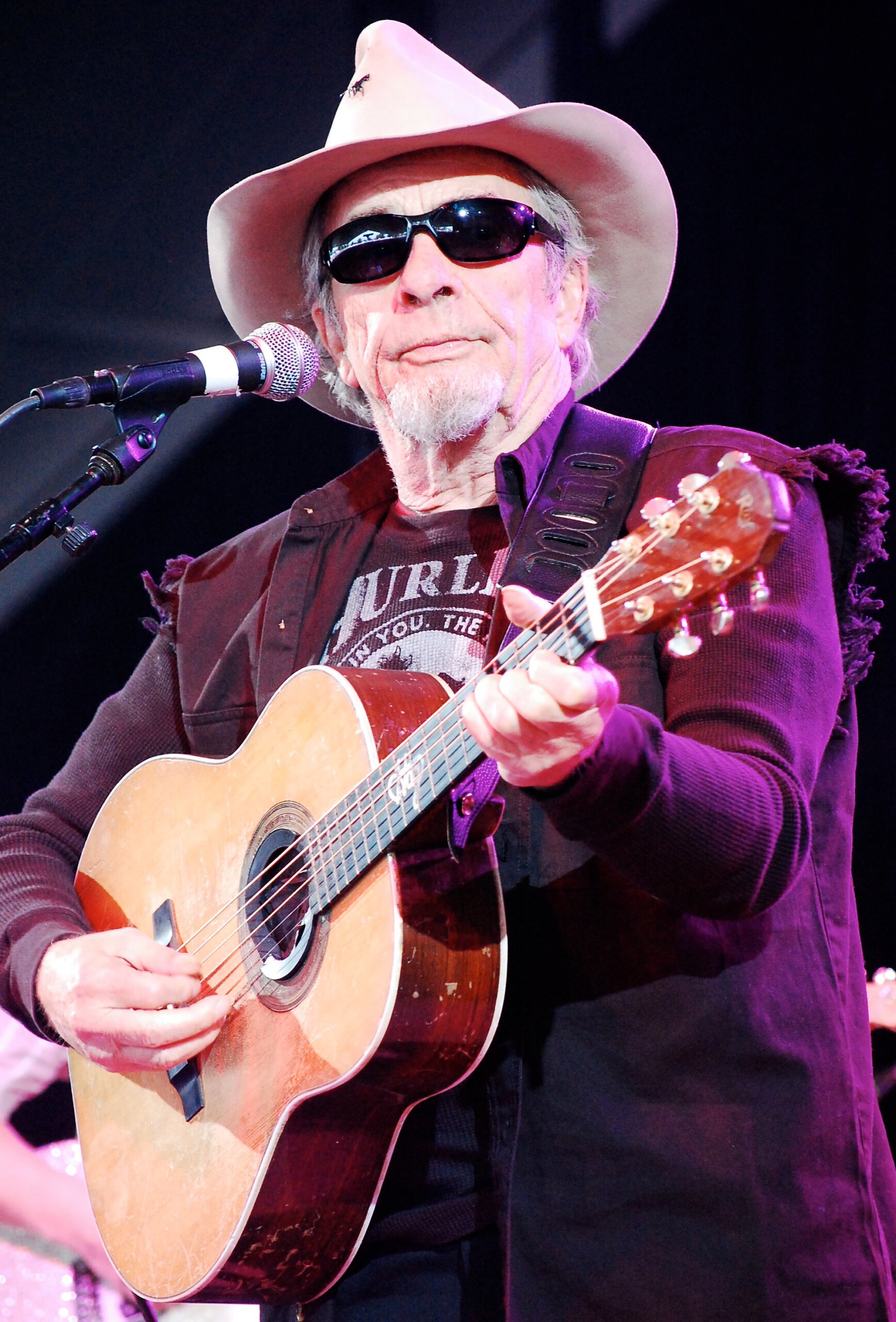Merle Haggard performing on stage in a white cowboy hat and sunglasses with his acoustic guitar, the songwriter behind Okie From Muskogee who later admitted he had smoked plenty of marijuana with Willie Nelson