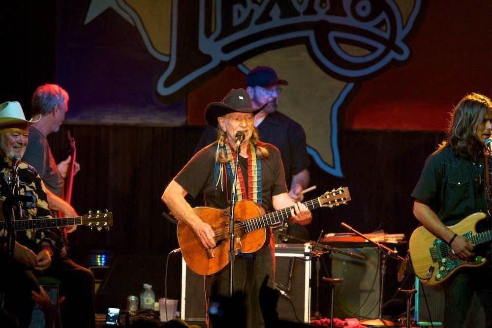 Willie Nelson with his guitar Trigger performing at the 4th of July Picnic in Texas under a giant Texas-shaped backdrop