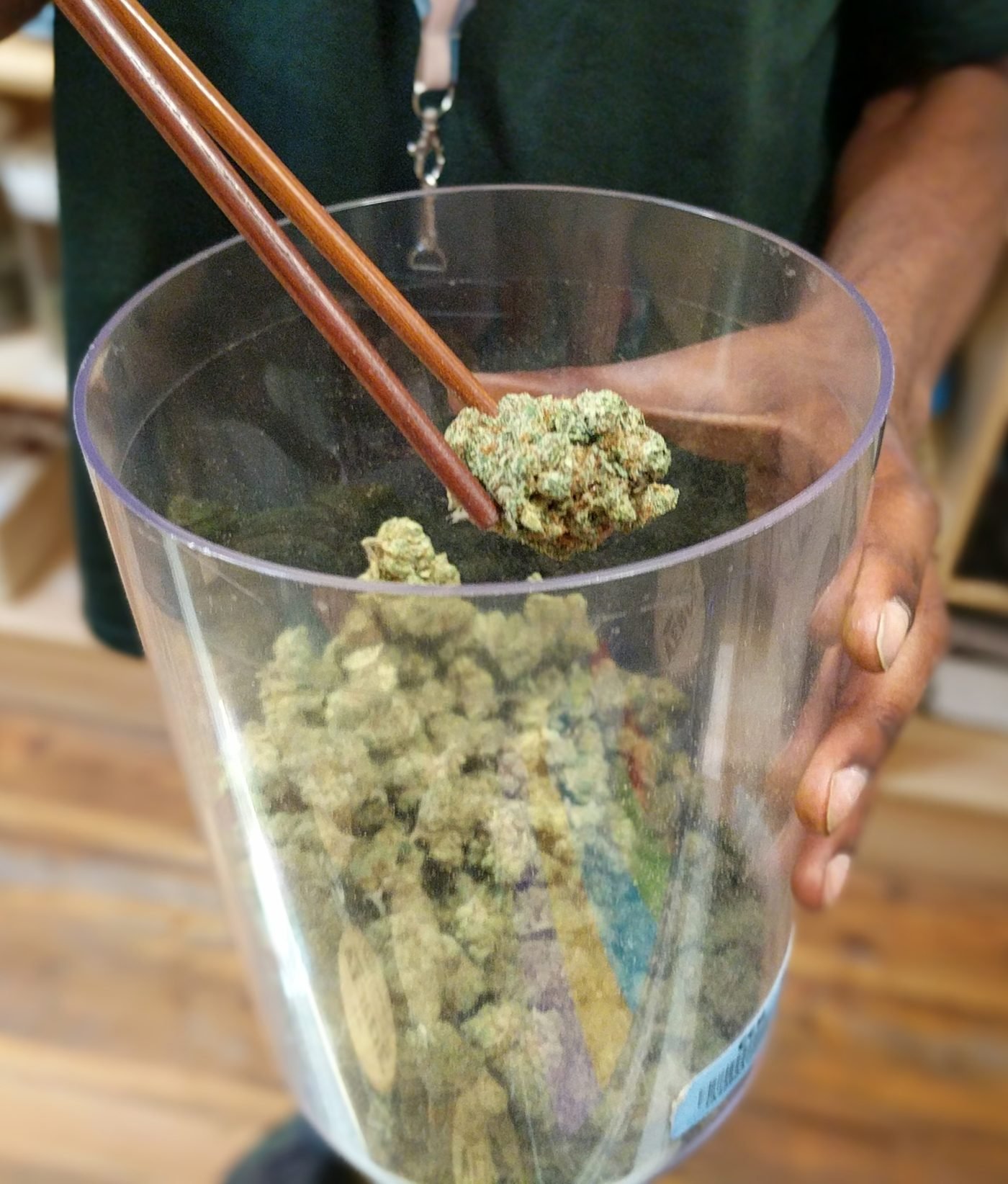 A budtender lifting a single cannabis bud with wooden tongs from a large clear display jar of flower at a high-volume recreational dispensary