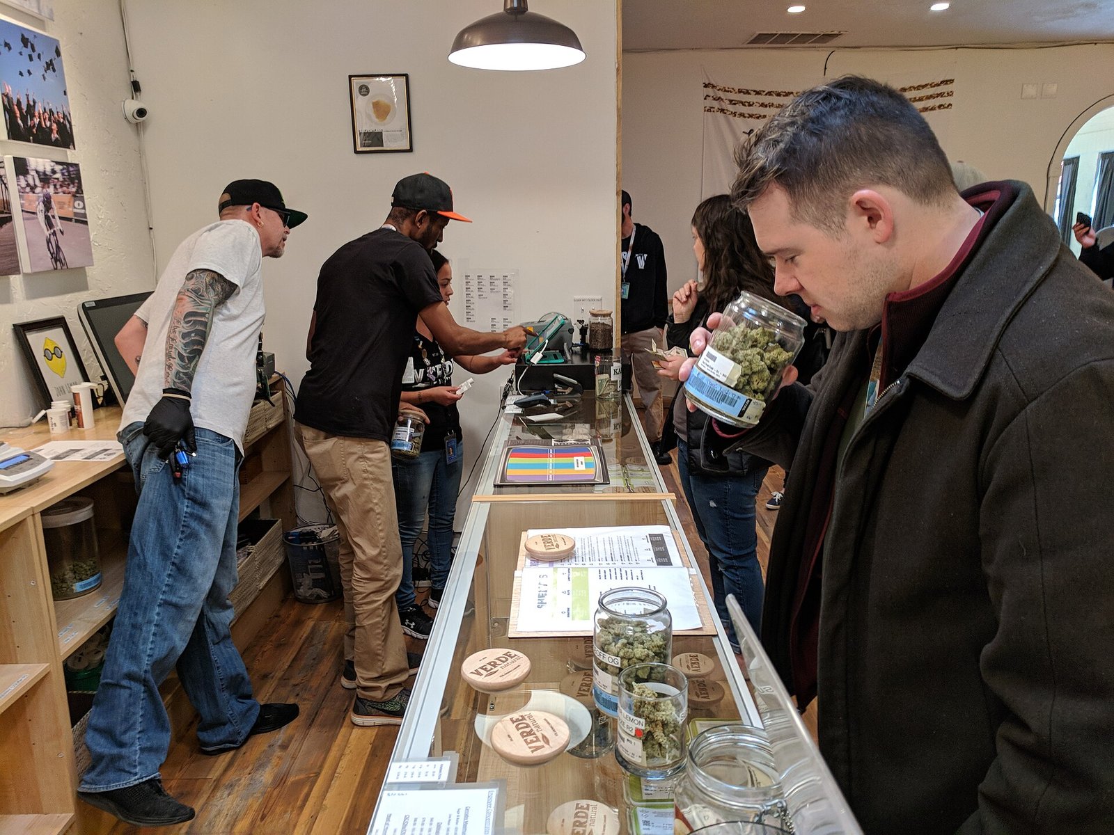 Customer smelling cannabis flower from a glass jar at a dispensary counter while two budtenders ring up the order