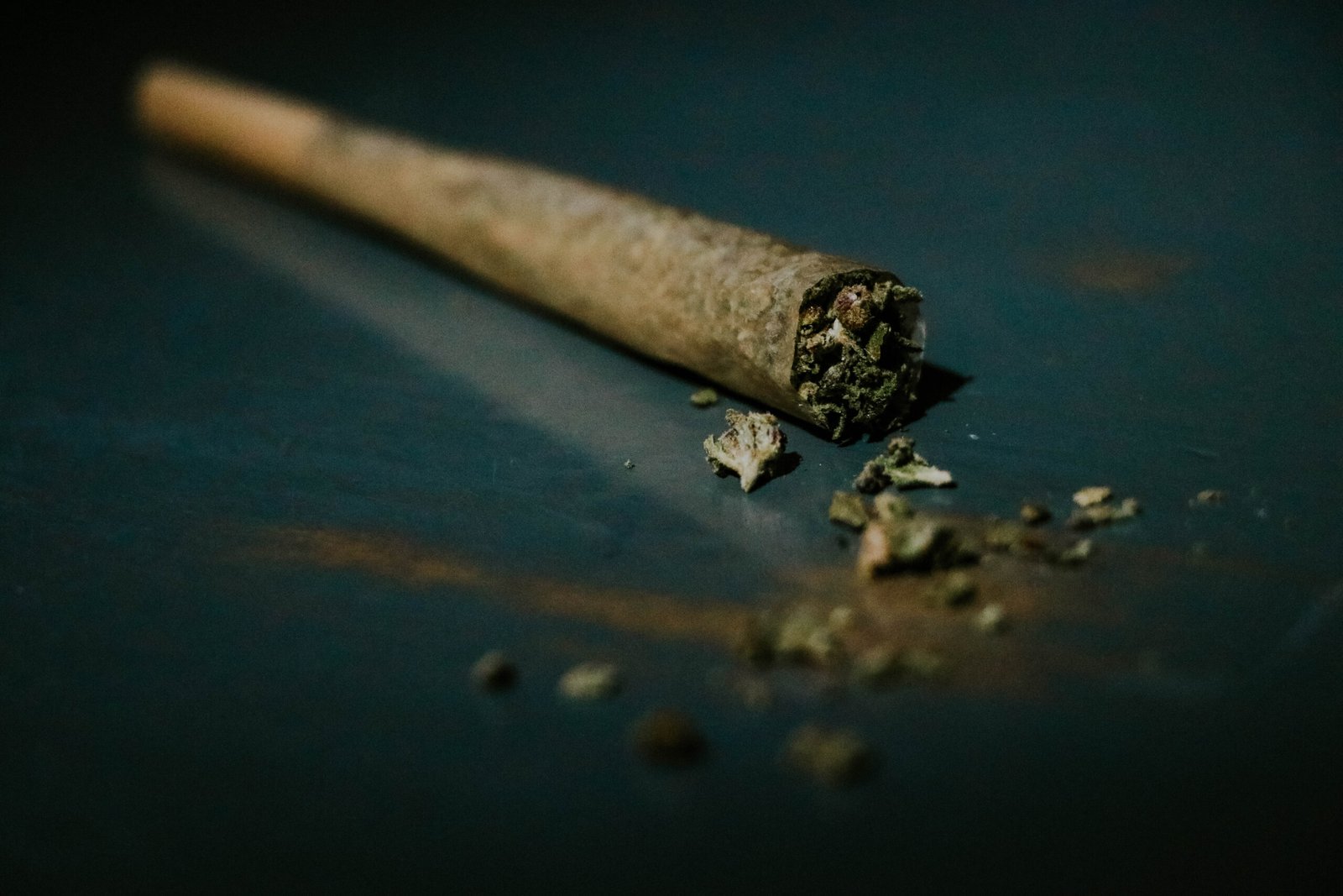 Lit pre-rolled joint resting on a dark wooden table with scattered loose cannabis flakes
