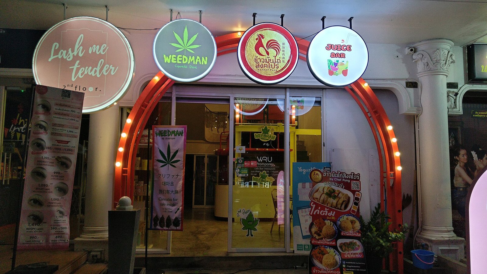 A shopfront in Bangkok with an illuminated Weedman cannabis store sign beside a lash salon and a juice bar